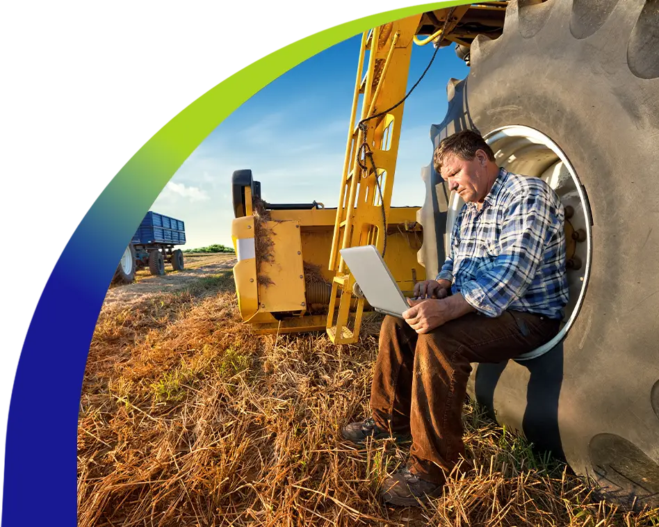 Farmer using a laptop while seated inside a tractor tire on a field, demonstrating the power of rural internet access for modern agriculture.