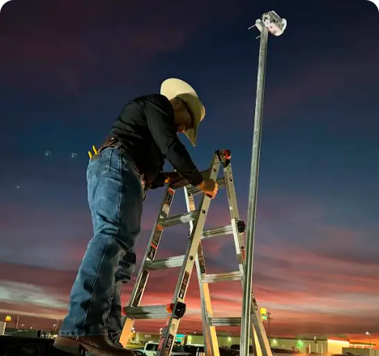 Technician in cowboy hat setting up a wireless broadband antenna at dusk using a ladder, with a dramatic sunset sky in the background.