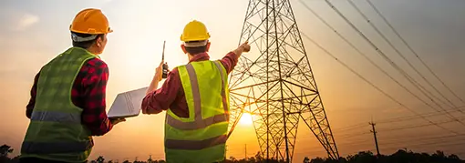 Two utility engineers inspecting a high-voltage transmission tower at sunset, using a laptop and two-way radio for field communication.